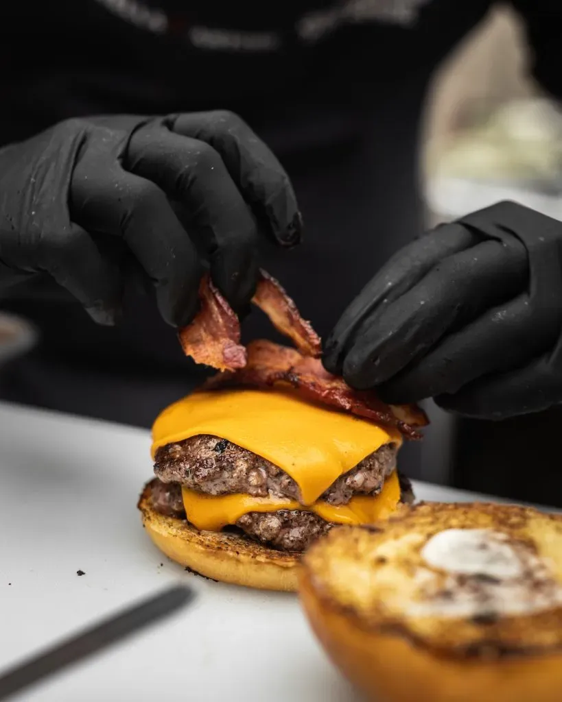 A close-up shot of a cook placing fried bacon over a slice of cheese while layering a burger