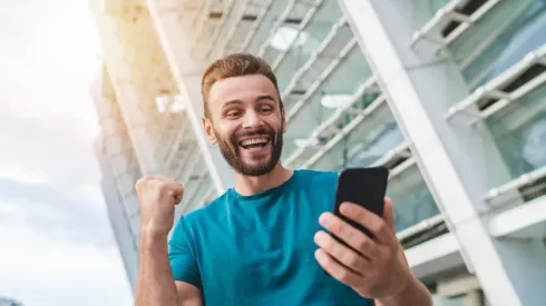 Handsome bearded man showing sincere excitement about his favorite team victory. Guy celebrating a bet win in online sport gambling application on his mobile phone clenching his fist in winner gesture
