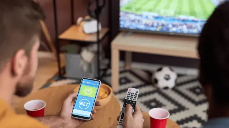 Imagem de mesa de madeira com vaso com chips e copos de plástico vermelho, bola de futebol e TV ao fundo, e imagem parcial de uma pessoa apostando ao vivo no celular