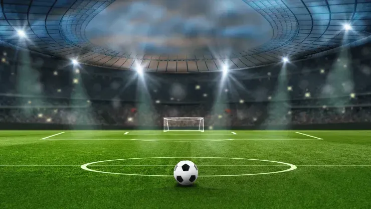 Bola de futebol junto do meio-campo, com baliza e arquibancada do estádio de fundo