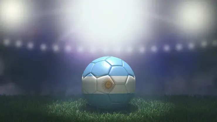 Bola de futebol com representação da bandeira da Argentina está sobre o gramado do campo, com luzes da arquibancada de fundo