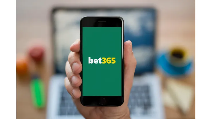 R06017 A man looks at his iPhone which displays the bet365 logo, while sat at his computer desk (Editorial use only).
