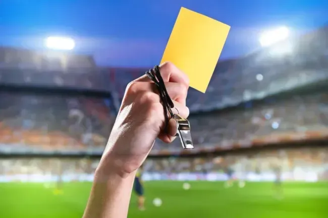 Imagem parcial de uma mão segurando um cartão amarelo e um apito, com a arquibancada de um estádio de futebol de fundo