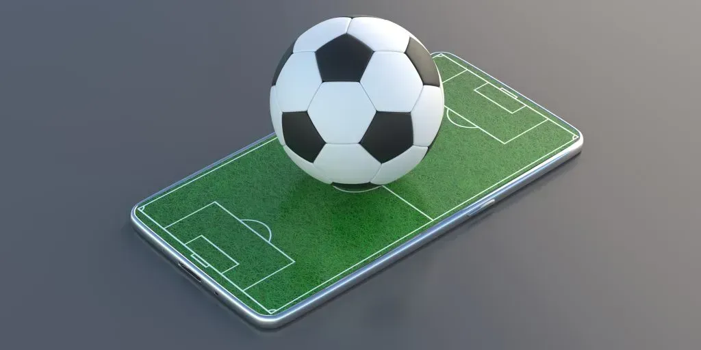 Fundo cinza sobre o qual se encontra um celular onde se vê um campo de futebol e uma bola 3D em cima da tela.