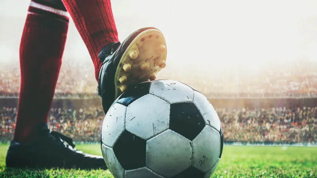 Pernas de jogador pisando na bola de futebol clássica, em branco e preto, com meião vermelho e chuteira preta. Ao fundo, estádio de futebol lotado fora de foco. 