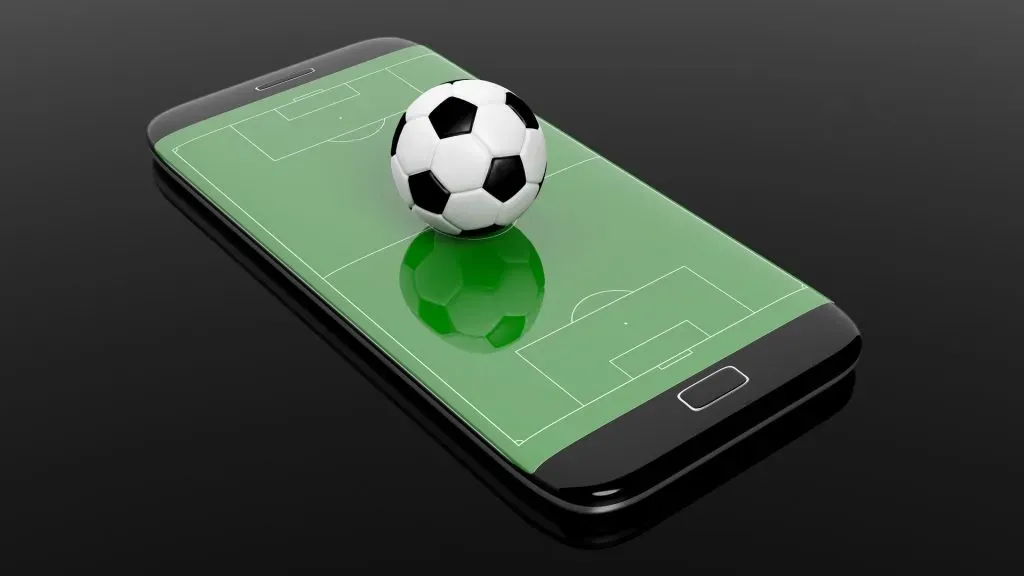 Smartphone com imagem de campo de futebol projetado na tela, com bola de futebol clássica, em branco e preto, em 3d, posando na superfície. 