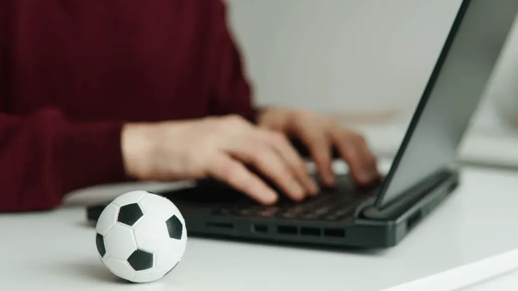 Foto de pessoa em notebook, usando suéter vermelho, com miniatura de bola de futebol clássica, em branco e preto, ao lado, simbolizando as apostas na EstrelaBet. 