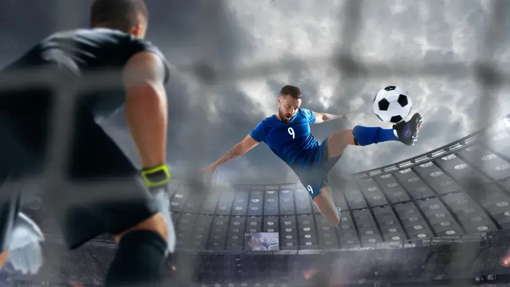 Jogador de futebol, equipado de azul, remata à baliza, com goleiro equipado de preto.