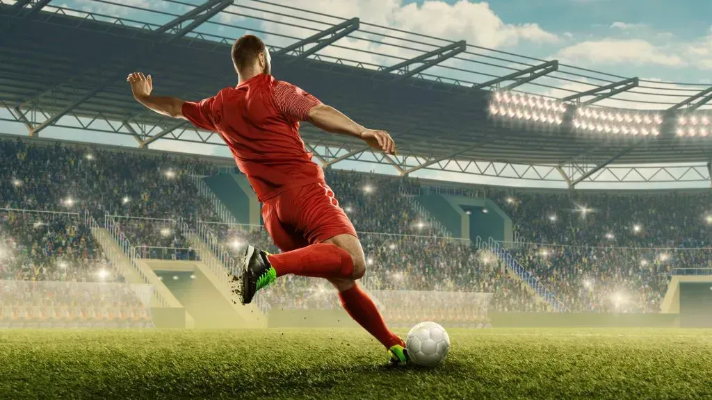 Jogador de futebol vestido de vermelho chuta bola do meio do campo, com bancada ao fundo