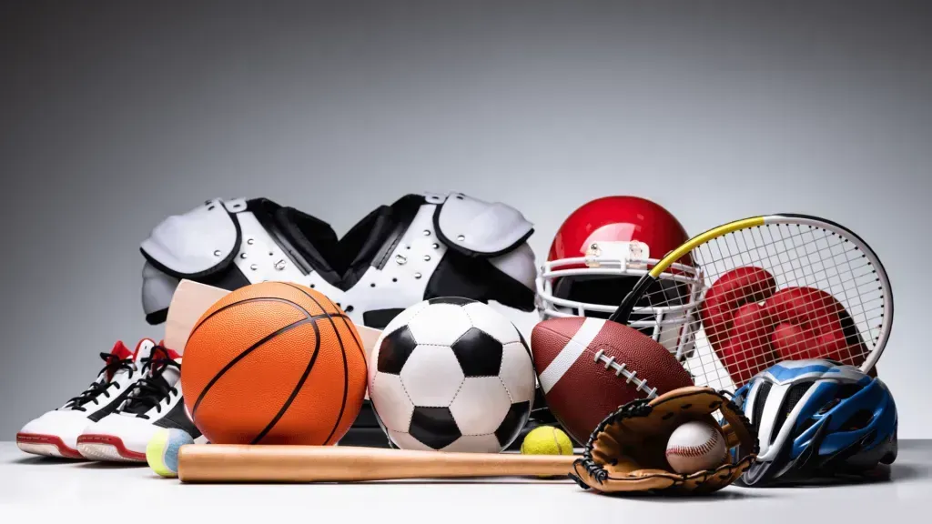 Imagem composta de variados elementos esportivos como sapato-tênis; bolas de basquete, futebol e rúgbi; proteção de ombros e capacete de futebol americano, raquete e bola de tênis, luvas de boxe; taco, luva e bola de beisebol; e capacete de ciclismo.