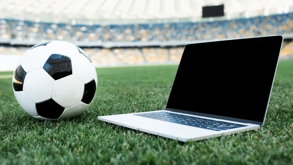 Imagem de bola de futebol clássica, em branco e preto, ao lado de notebook, ambos no gramado de campo de futebol. 