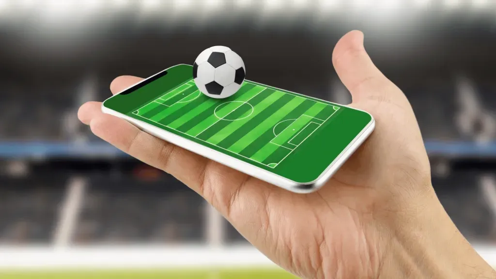 Imagem gráfica de mão segurando smartphone com campo de futebol projetada na tela. Em cima da tela, uma mini bola de futebol clássica, em branco e preto. 
