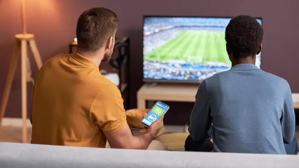 Dois amigos lado a lado no sofá, um deles com smartphone em mãos com casa de apostas aberta na tela. Ao fundo, TV ligada com jogo de futebol passando. 