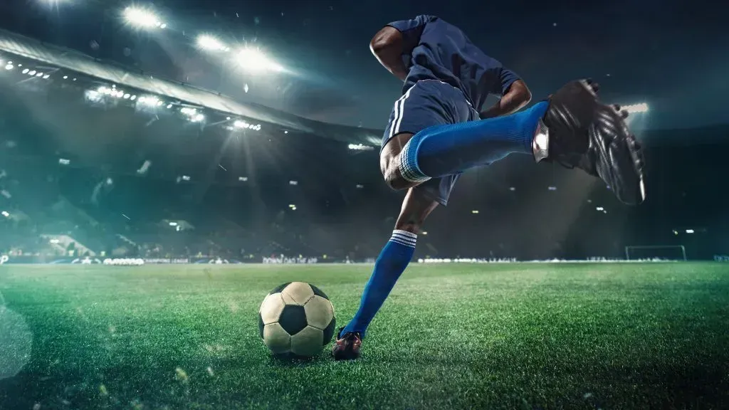 Jogador de futebol equipado de azul, está de costas e prestes a chutar a bola para o meio do campo