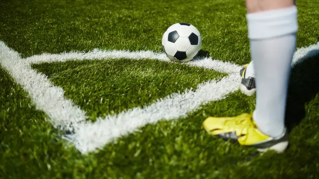 Pequena bola de futebol pousada no canto do campo de futebol, e à esquerda pernas de jogador com meias brancas e chuteiras amarelas