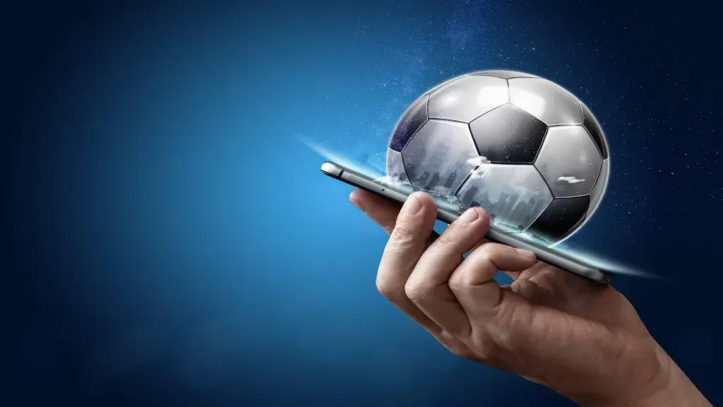 Mão segurando smartphone contra fundo azul. Há uma bola de futebol sendo projetada para fora da tela.