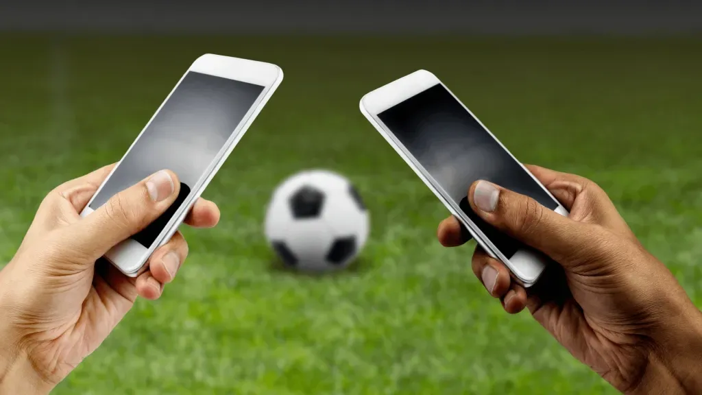 Duas mãos, de pessoas distintas, segurando dois smartphones. Ao fundo, bola de futebol clássica, em branco e preto, em gramado de campo de futebol. 