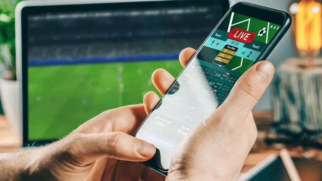 Mão segurando smartphone com casa de apostas aberta na tela. Ao fundo, TV ligada em jogo de futebol. 