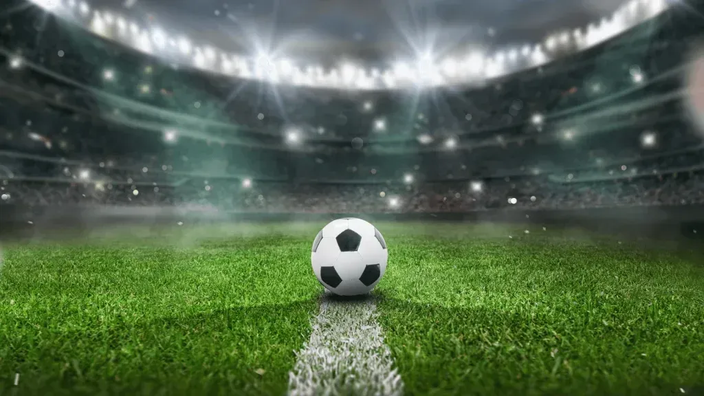 Clássica bola de futebol branca e preta posicionada no centro de um estádio, acima de uma das linhas brancas que demarcam o campo. 