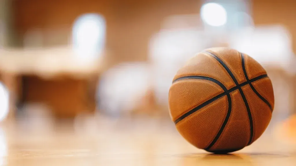 Foto de bola de basquete em foco, do lado direito, com fundo em tons de branco, marrom e laranja desfocado, representando os melhores sites de apostas na NBA. 