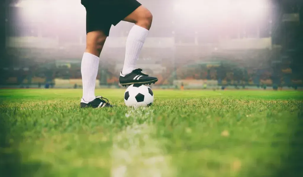 Atleta com chuteiras pretas e meias brancas apoia pé sobre bola de futebol, com campo e arquibancadas de fundo