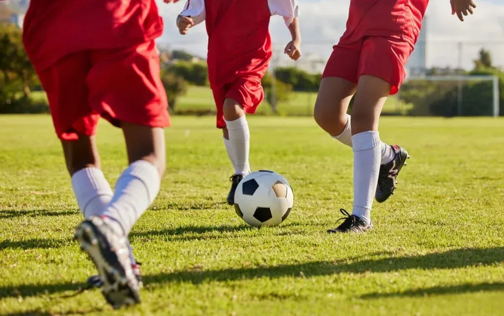 Três jogadores com equipagens em tons de vermelho e branco jogando em campo. Ao centro, bola de futebol clássica, em branco e preto.