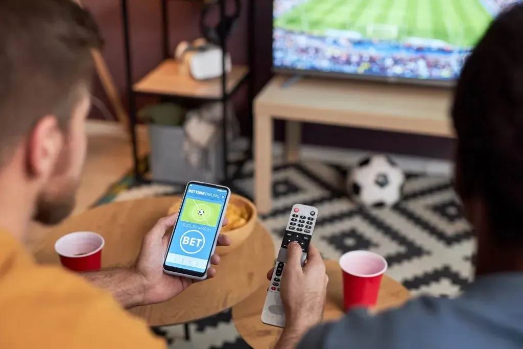 Imagem de dois amigos, lado a lado, assistindo jogo de futebol na TV em sala de estar. Um deles segura smartphone aberto em casa de apostas com a mão esquerda e controle remoto com a direita. 