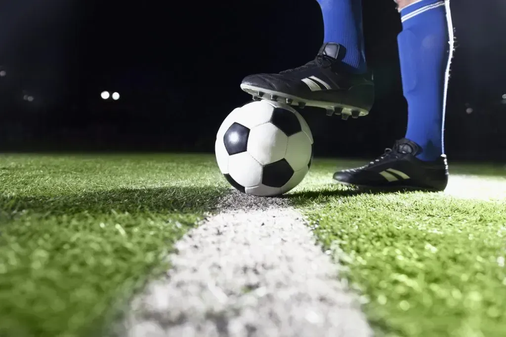 Imagem parcial de um jogador de futebol no gramado, com grande plano de chuteiras pretas, meias azuis e bola de futebol sobre a linha de campo