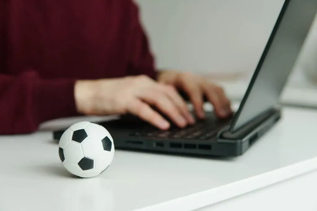 Pessoa usando notebook, apenas torso e mãos à vista, com bola de futebol clássica, em branco e preto, ao lado esquerdo na mesa. 