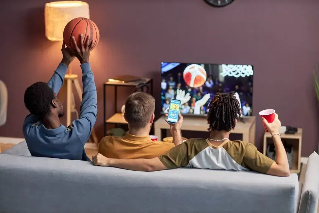 Três homens de costas, sentados no sofá, assistem uma partida de basquete na TV. O da esquerda veste uma camisa azul e segura uma bola de basquete, o do meio usa um polo amarelo e segura um celular que diz 