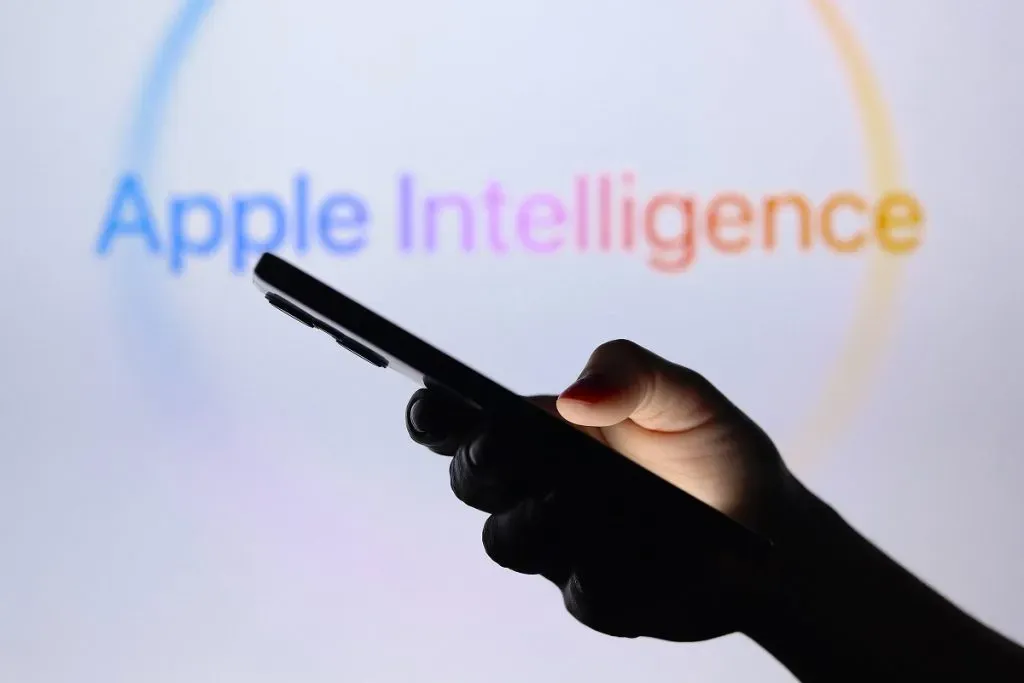 Ilustración del logotipo de Apple Intelligence visto en el fondo con una mano silueta sosteniendo un iPhone | Foto: Imago