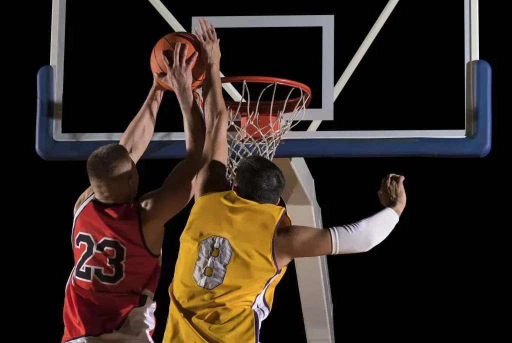 Dois jogadores de basquete disputam bola junto da tabela, o da esquerda equipado de vermelho e o da direita usando amarelo