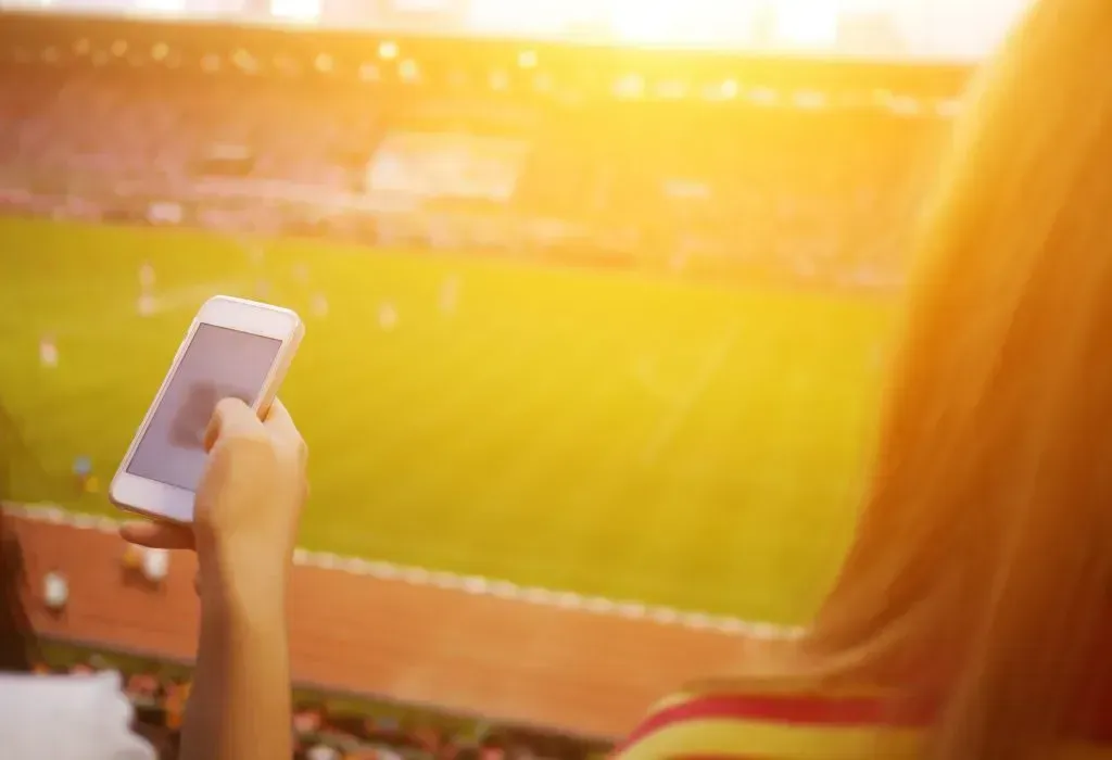 Imagem parcial de uma mulher na bancada segurando um iPhone, com o campo de futebol mais abaixo