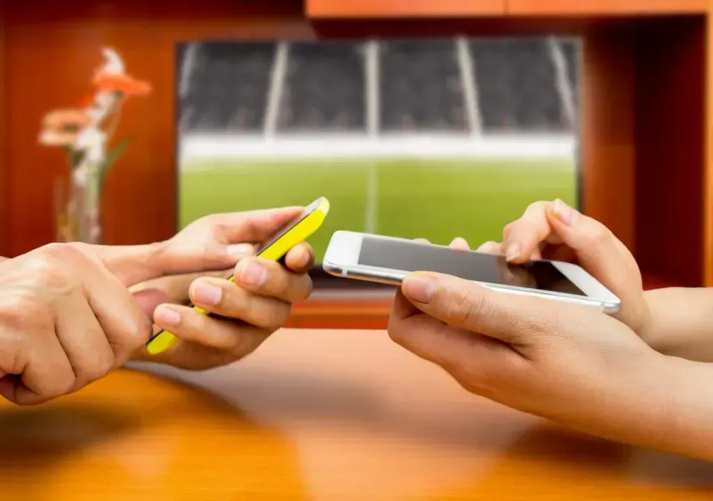 Mãos de duas pessoas seguram um celular branco e um amarelo, com tela desligada, e uma TV de fundo mostra um campo de futebol 