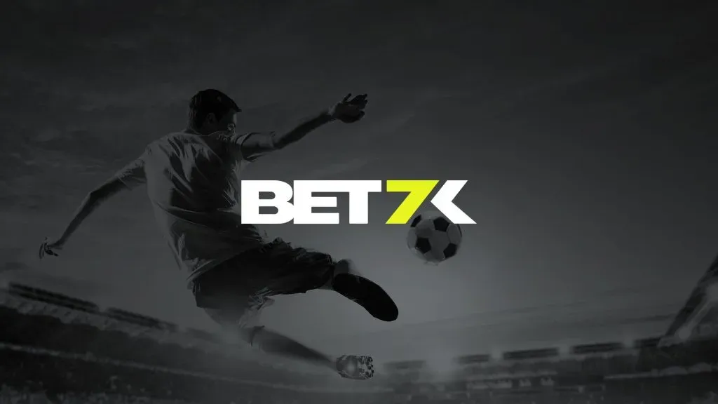 A imagem é um banner para a Bet7k. Ela mostra um jogador de futebol no ar, prestes a chutar uma bola em um estádio cheio de pessoas. A imagem está em preto e branco e diz Bet7k no meio.