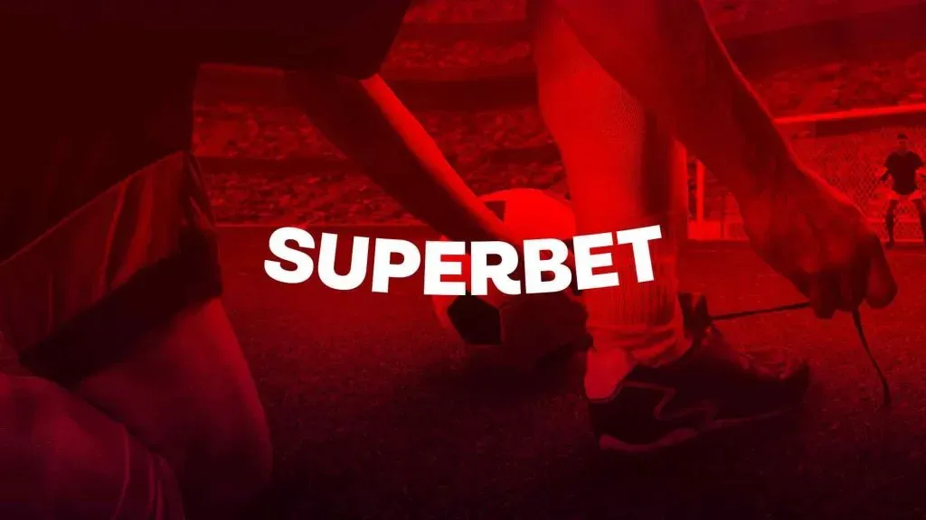 A imagem é um banner para a Superbet e mostra um jogador de futebol amarrando os cadarços no meio de um campo de futebol, ao lado de uma bola de futebol. A imagem tem um filtro vermelho.