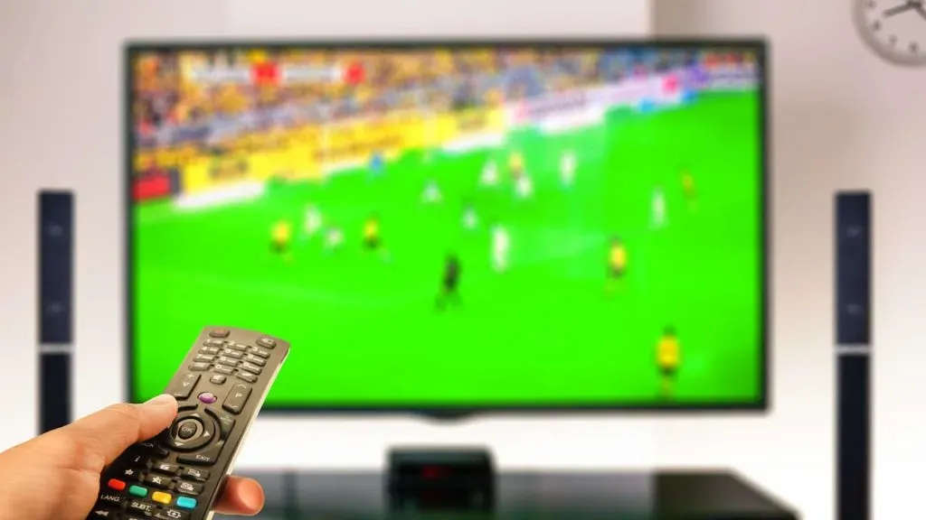 A imagem apresenta uma TV com um jogo de futebol da Copa América desfocado e uma mão com um controle remoto controlando a TV.