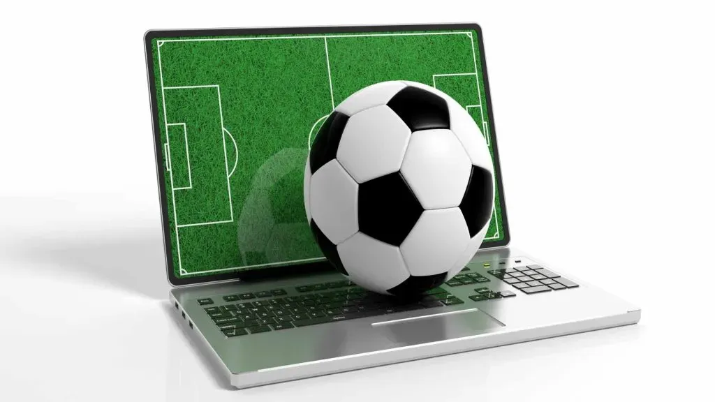 A imagem mostra um laptop com uma bola de futebol preta e branca em cima do teclado e um campo de futebol como protetor de tela.