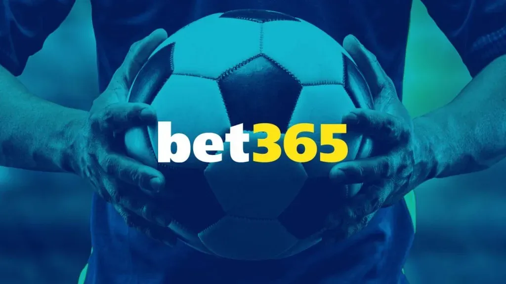 A imagem é um banner para a bet365. Diz bet365 no centro e o fundo mostra um jogador de futebol segurando uma bola de futebol preta e branca nas mãos. A imagem de fundo também tem um filtro azul.