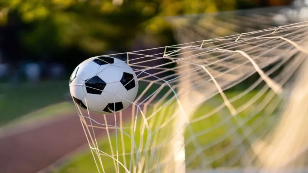 A imagem mostra uma bola de futebol preta e branca empurrando a rede do gol para trás, indicando que alguém marcou um gol.
