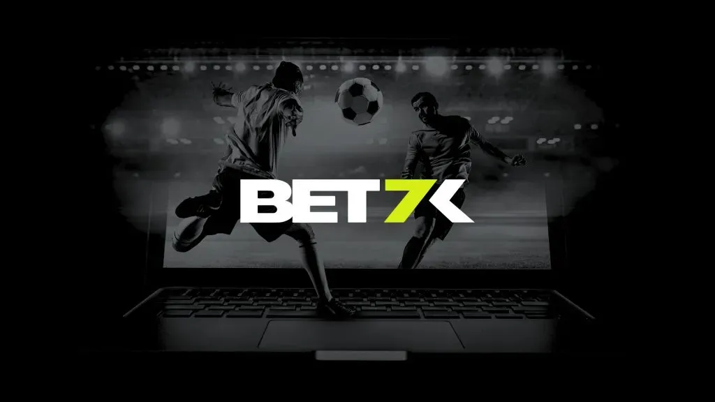A imagem é um banner para a Bet7k. Diz Bet7k no centro. O fundo é preto e branco e mostra dois jogadores de futebol prestes a chutar uma bola que está no ar. Os jogadores estão em cima do teclado de um laptop.