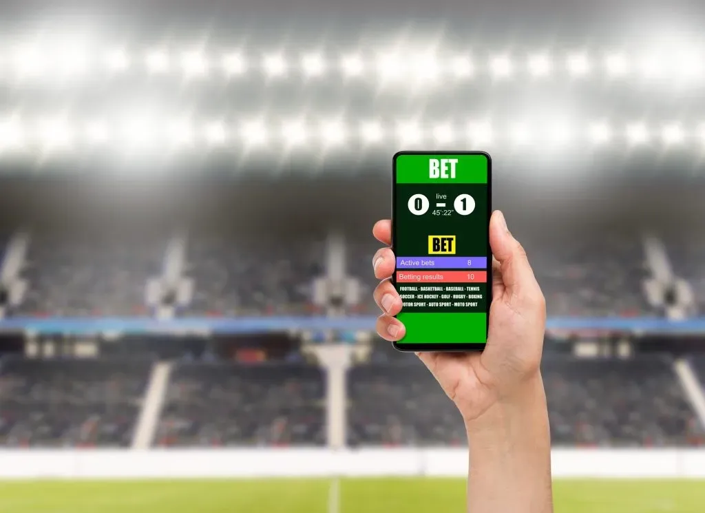 Mão suspendendo smartphone aberto em app de apostas. Ao fundo, pode-se ver as luzes e arquibancadas de um estádio lotado para partida de futebol. Na tela do smartphone, pode-se ver indicado o resultado de uma partida,