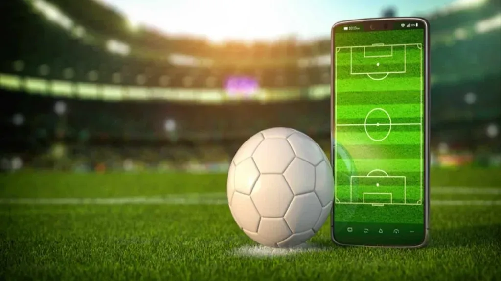 Imagem gráfica de bola de futebol branca ao lado de smartphone em estádio de futebol.