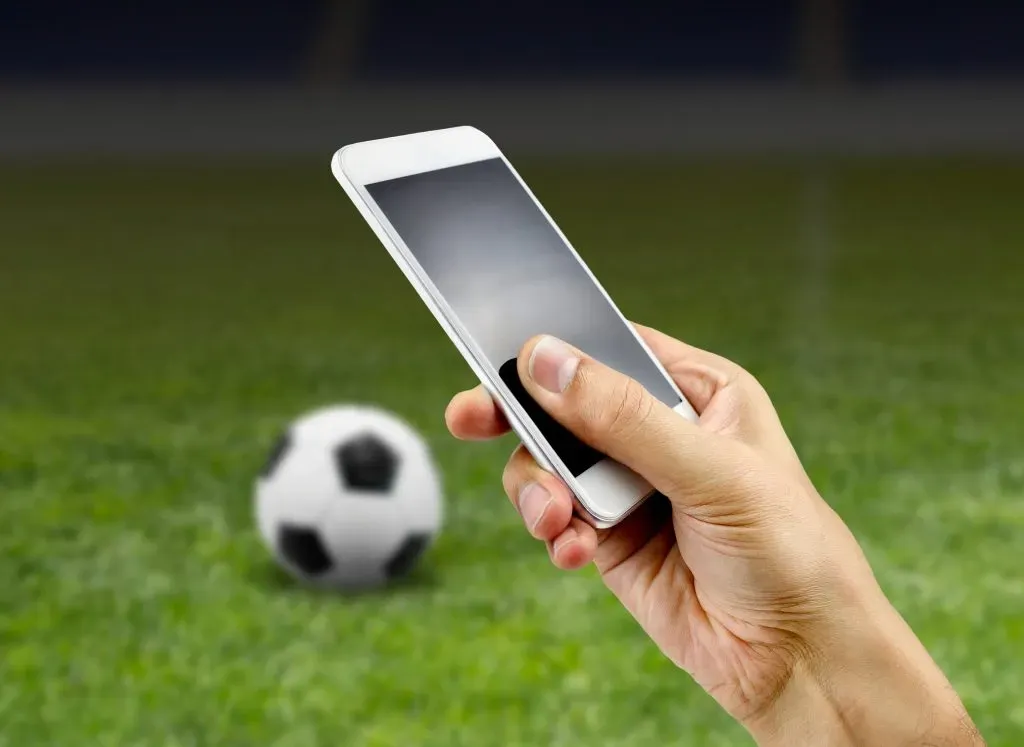 Mão segurando smartphone. Ao fundo, bola de futebol clássica, em branco e preto, em gramado de campo de futebol.