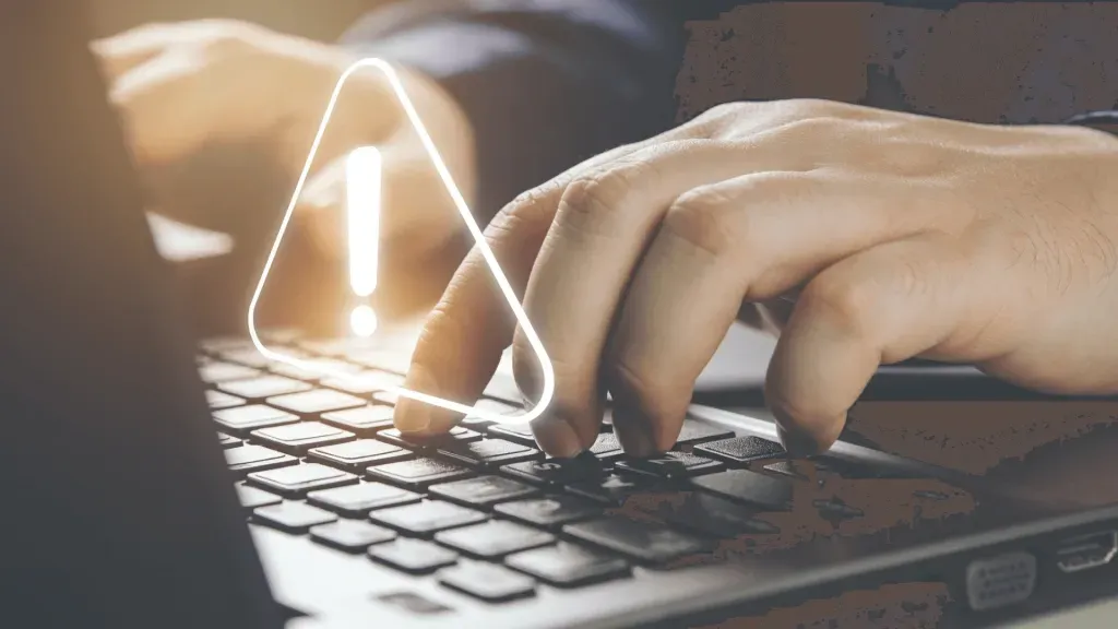 Pessoa manuseando notebooks, apenas as mãos à vista, com sinal de alerta, no formato de triângulo com ponto de exclamação ao centro, flutuando logo acima do teclado.