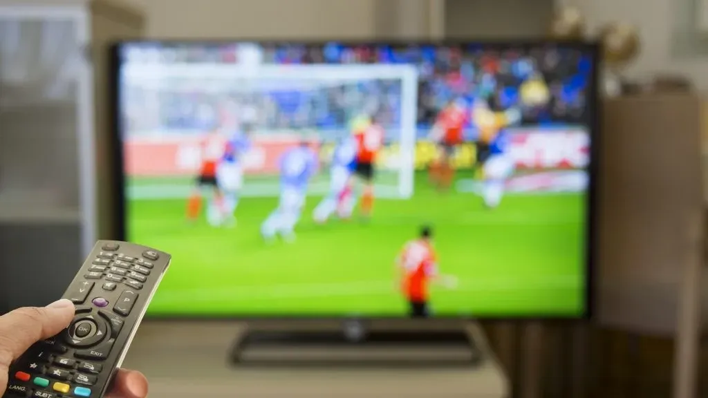 Foto de mão apontando controle remoto para TV ligada em jogo de futebol.