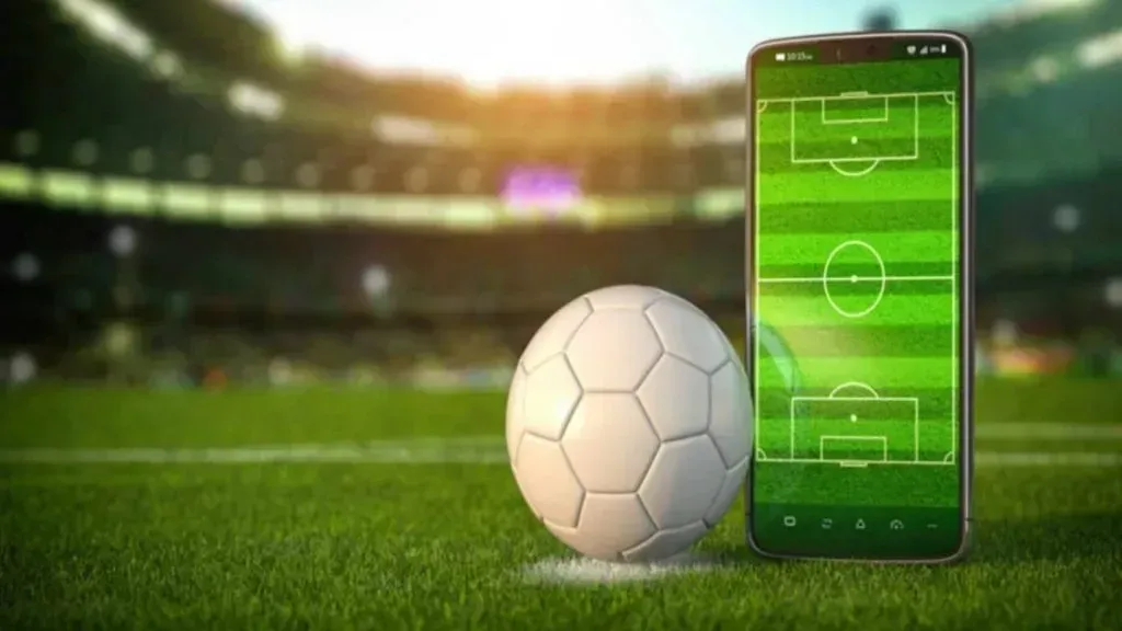 Uma bola branca de futebol e um celular com a tela representando um campo de futebol estão sobre o gramado do estádio