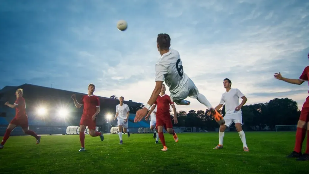 Jogadores em campo, com bola ao ar, observando enquanto um deles se prepara para fazer um voleio.