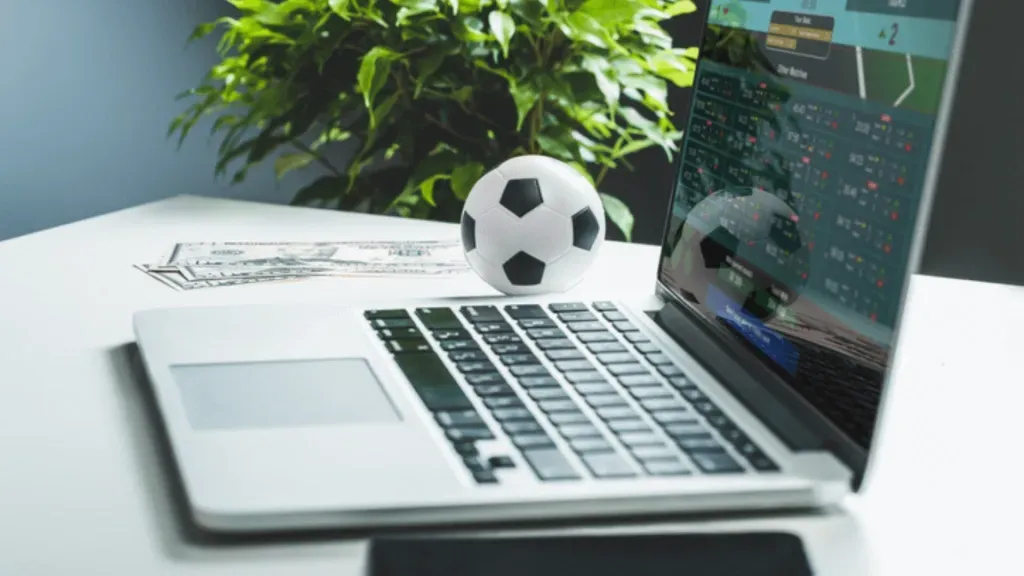 Notebook com bola de futebol clássica do seu lado esquerdo, junto de algumas notas de dinheiro.