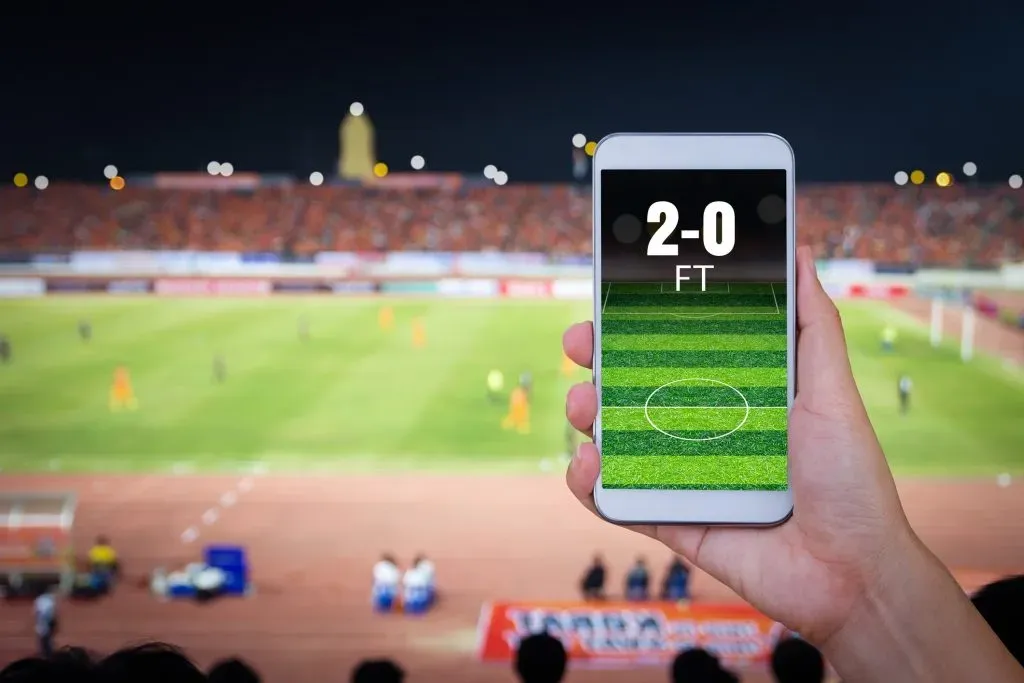 Smartphone sustendido por mão contra fundo de partida de futebol em estádio. Na tela, pode-se ler